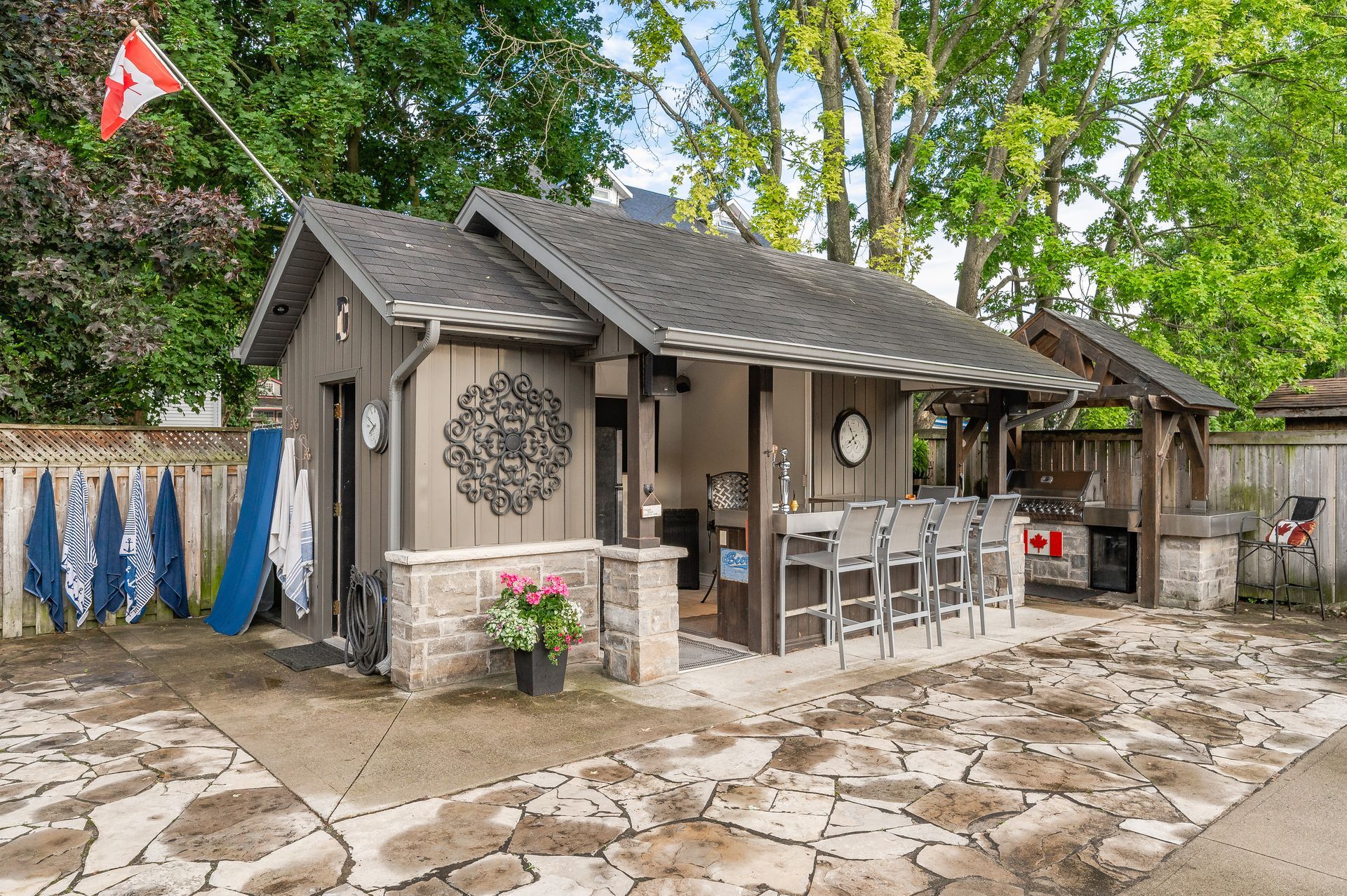 Rustic stone patio with a small outdoor cabin, dining table, chairs, and flower planter under trees