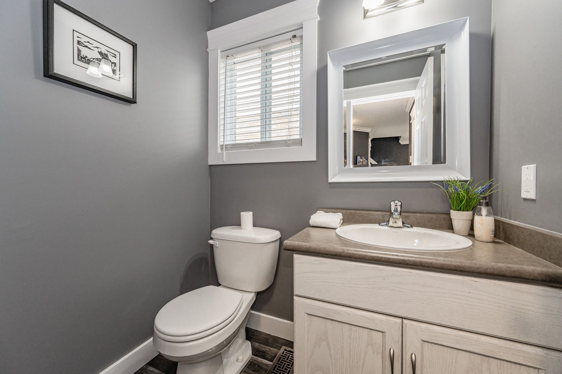 Small gray bathroom with toilet, sink vanity, mirror cabinet, and window with blinds.