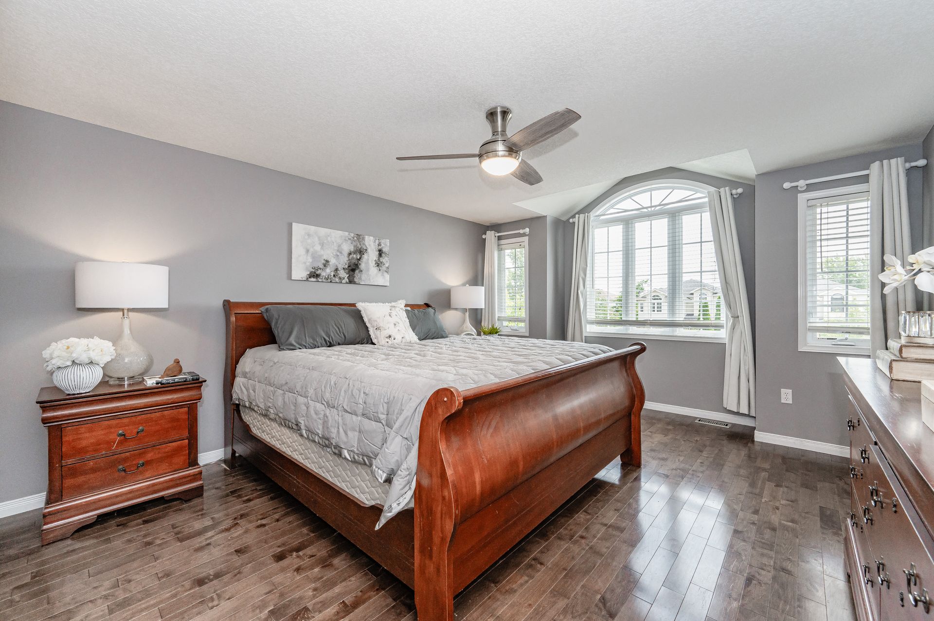 Spacious gray bedroom with wooden bed, ceiling fan, and large arched windows.