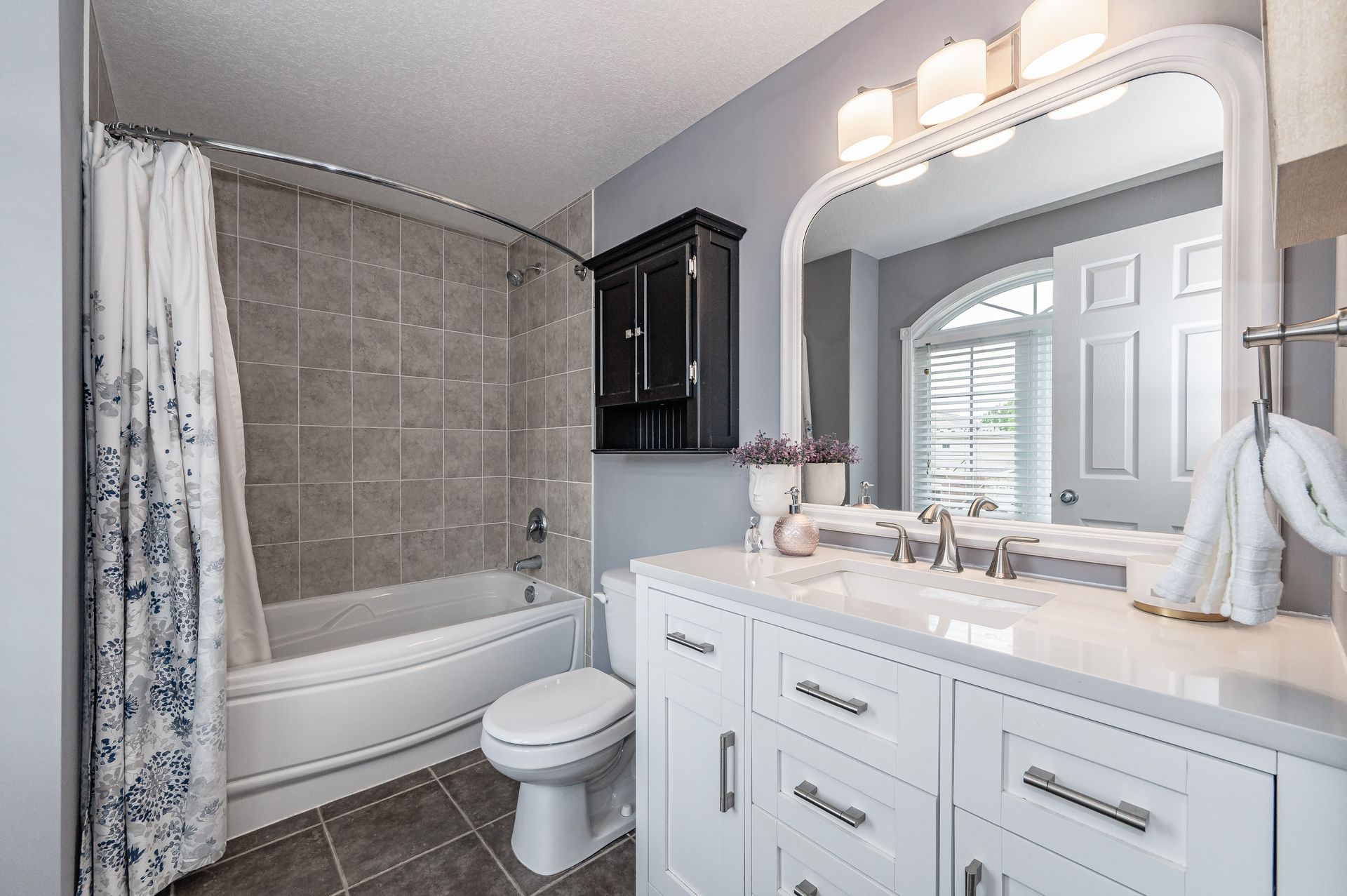 Bright bathroom with white vanity, large mirror, tub-shower, and gray tiled walls