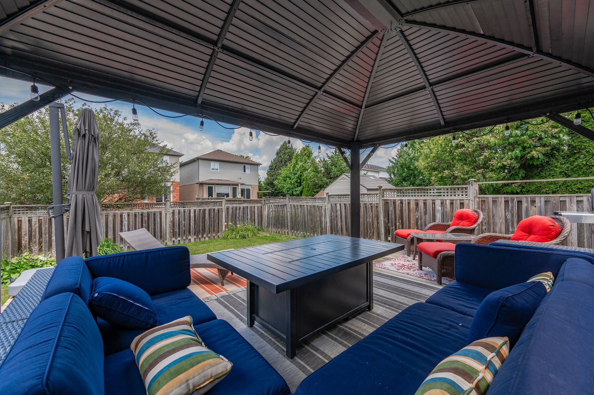 Covered backyard patio with blue seating, red chairs, and a central fire pit under a pergola.