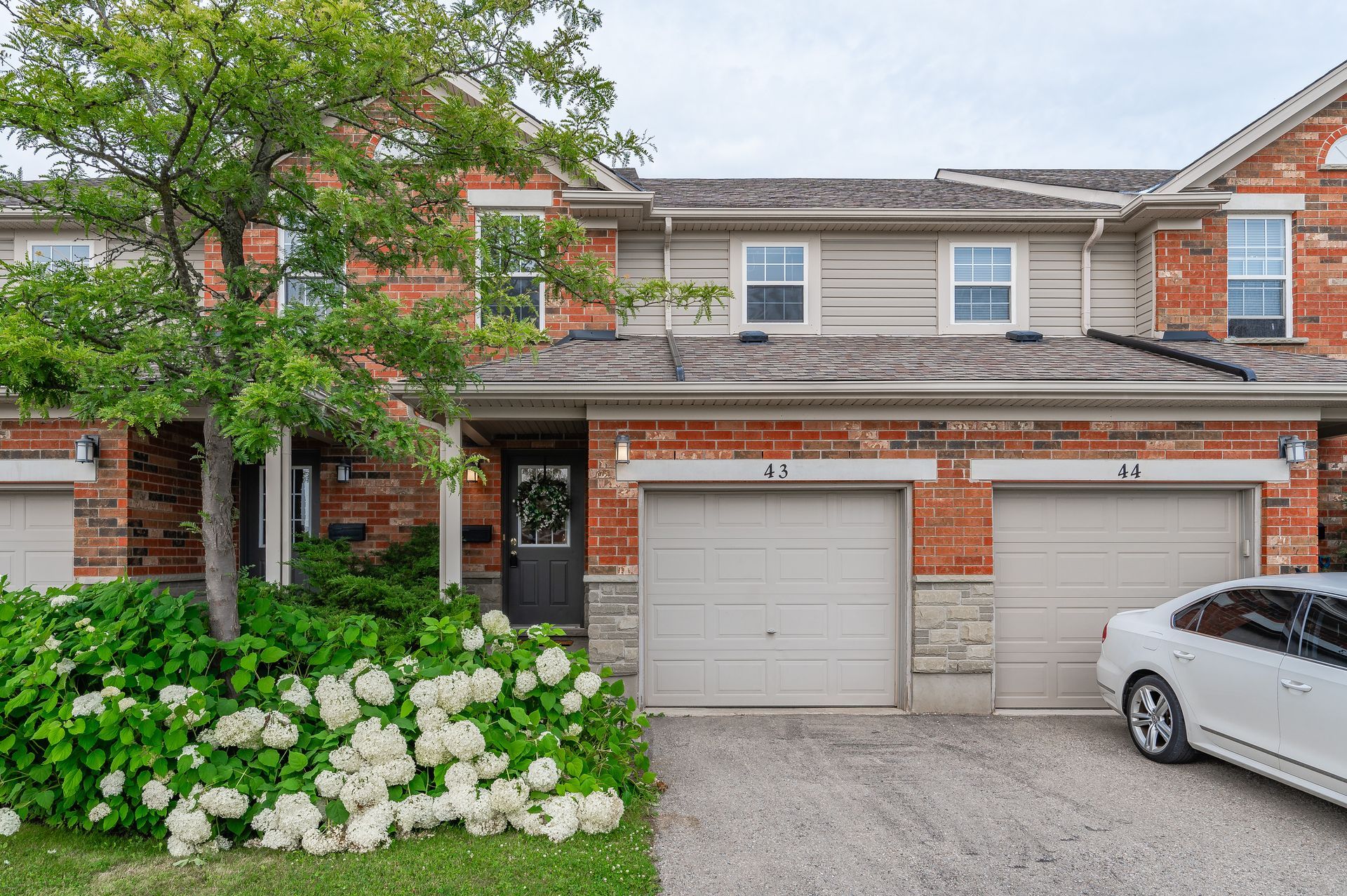 Townhouse with two garage doors, brick and beige siding, white flowers, and a silver car parked outside