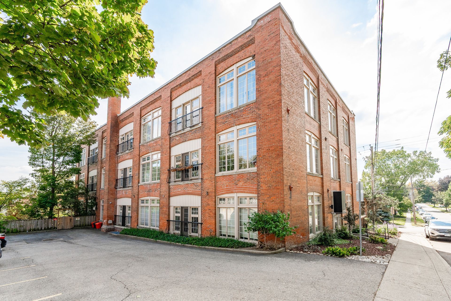 Three-story red brick building with large windows, surrounded by trees and a gravel lot