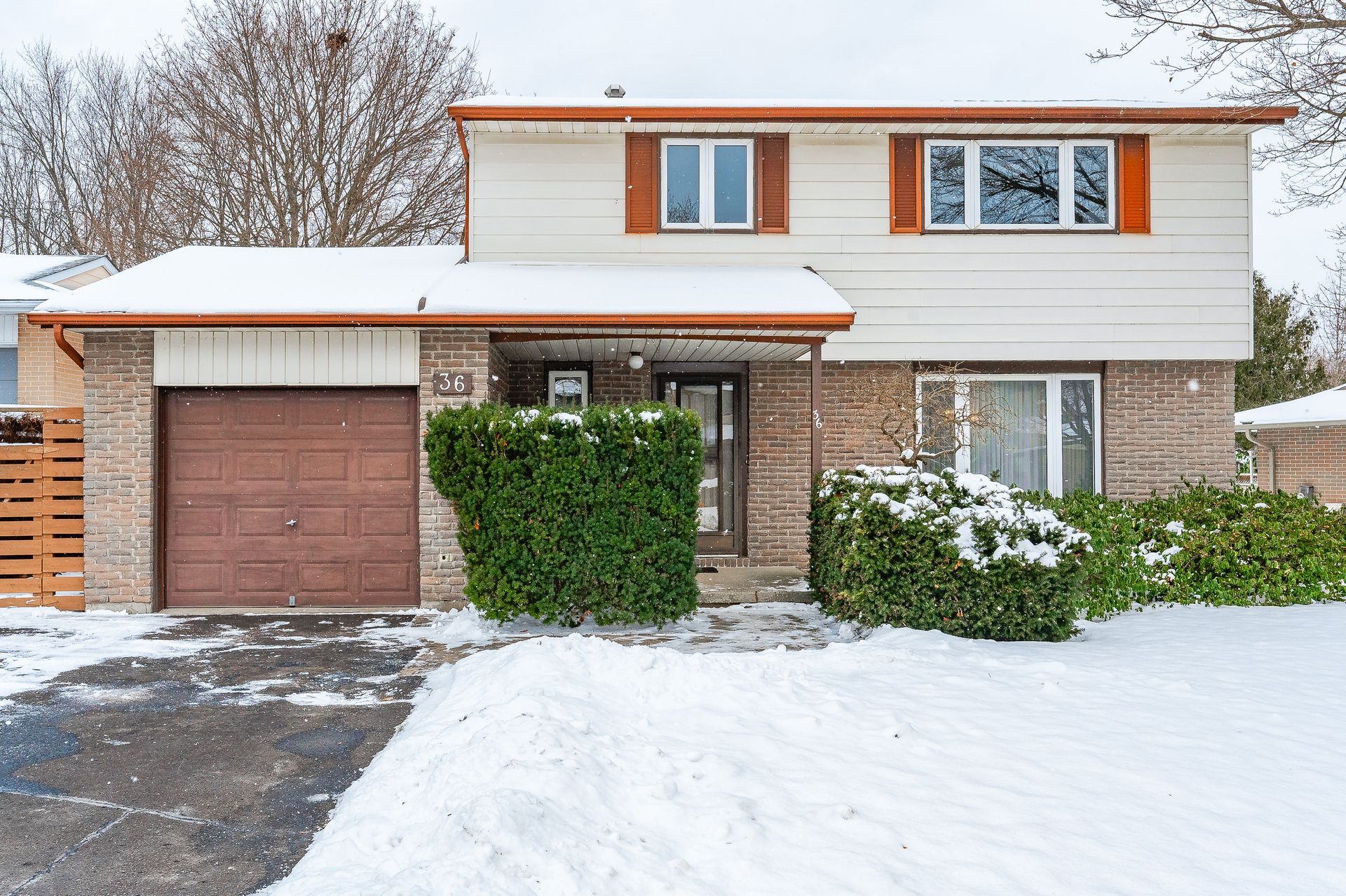 Snow-covered suburban two-story house with brown garage and shutters, front bushes, and driveway