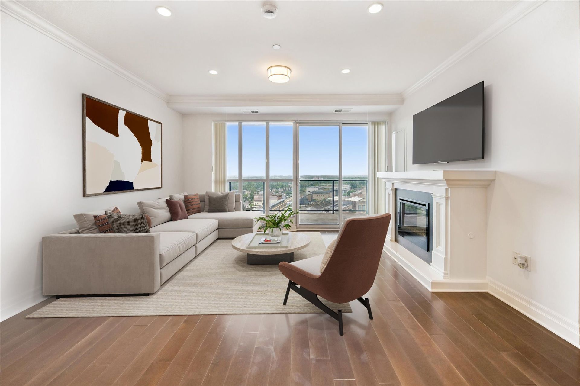 Bright living room with sectional sofa, lounge chair, fireplace, wall-mounted TV, and city-view windows