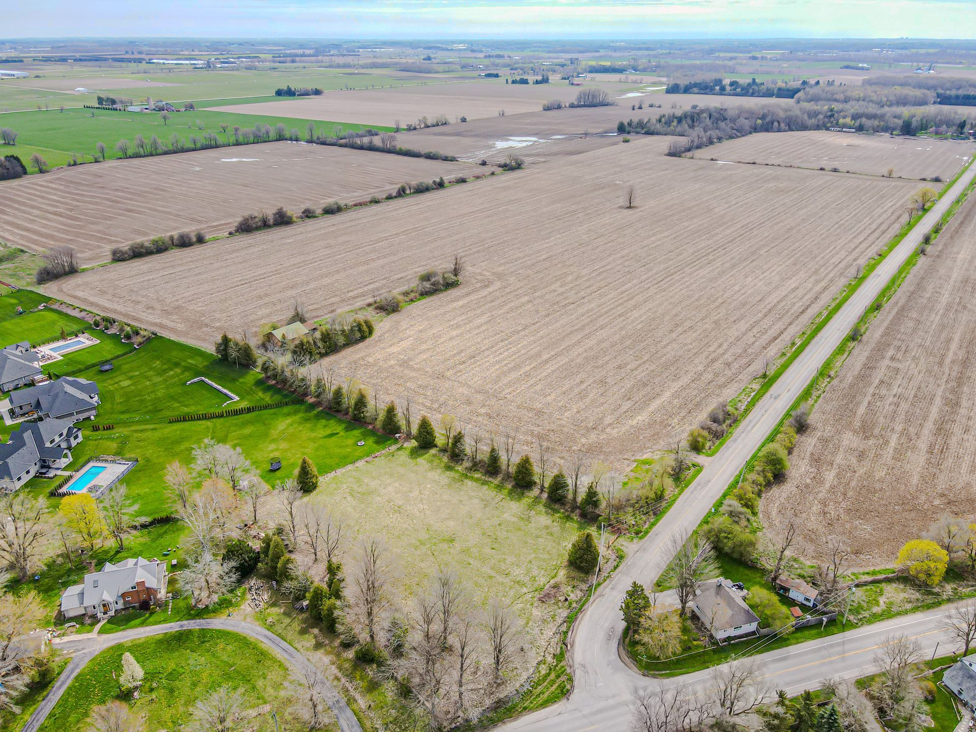 Aerial view of farmland with large tan fields, a tree-lined road, and scattered houses on a cloudy day