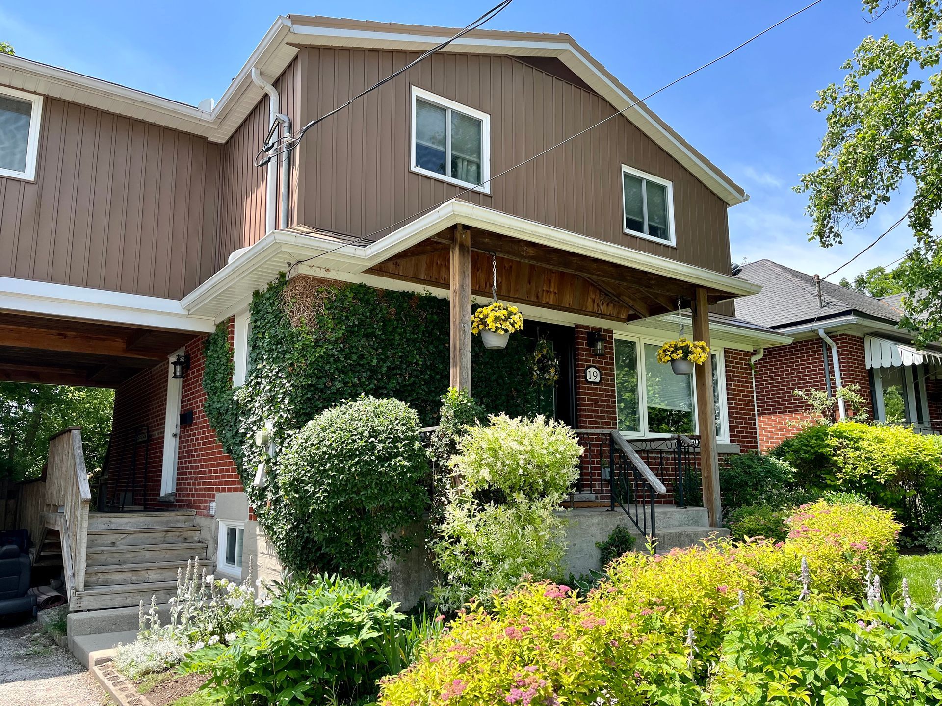 Brown house with ivy-covered front porch, green shrubs, and steps along a sunny residential street