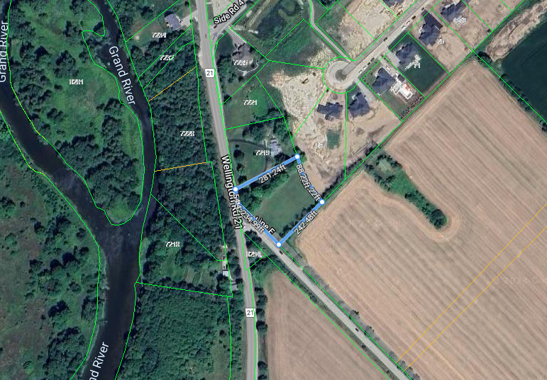 Aerial view of a rural road beside green trees and fields, with a small cluster of buildings.