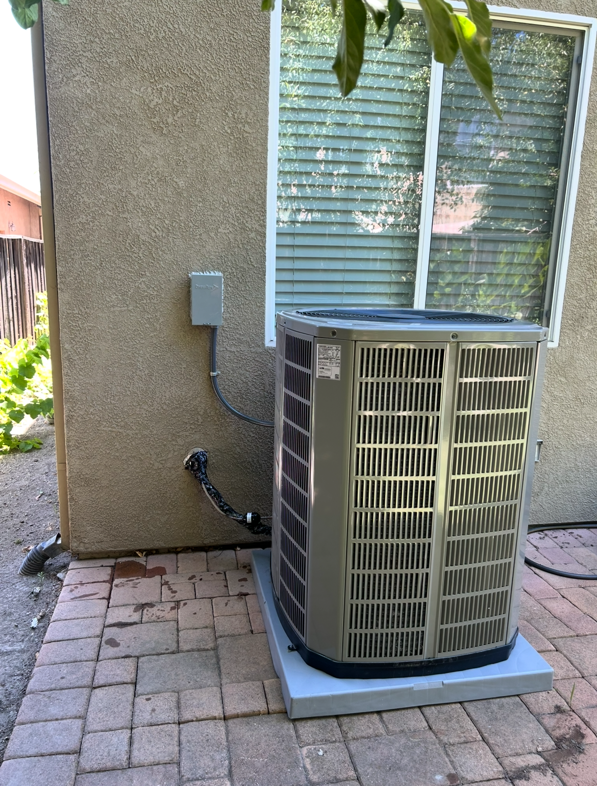 Outdoor HVAC unit beside a stucco house wall and window, on a brick patio.