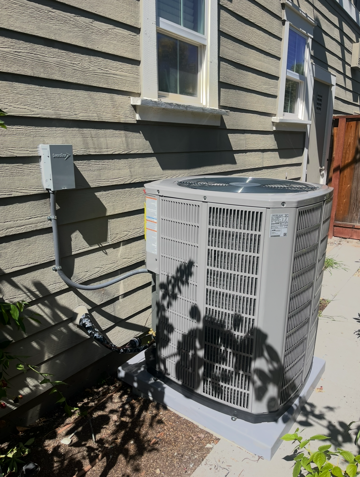 Outdoor air conditioner unit beside a beige house wall, partly shaded by tree leaves.
