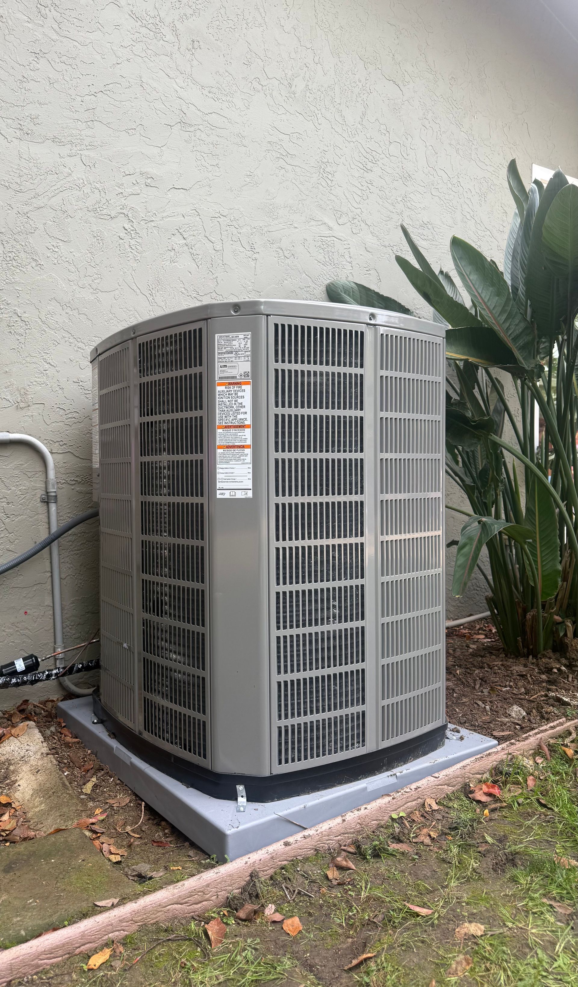 Outdoor HVAC unit beside a stucco wall, on a concrete pad next to plants and grass.