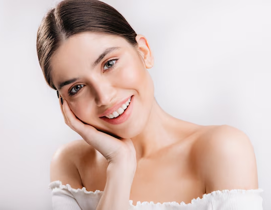 Woman smiling, touching cheek, white off-the-shoulder top, soft lighting.