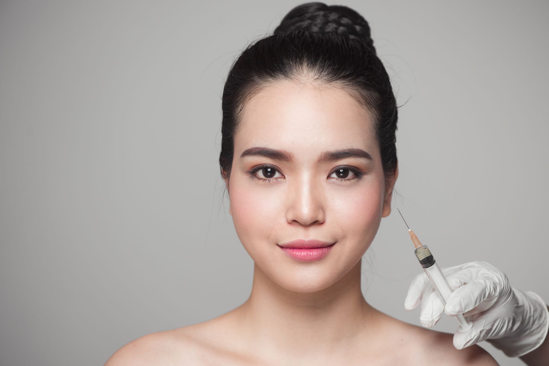 Close-up of a woman facing forward while a gloved hand holds a syringe near her cheek. Close-up of a woman facing forward while a gloved hand holds a syringe near her cheek.