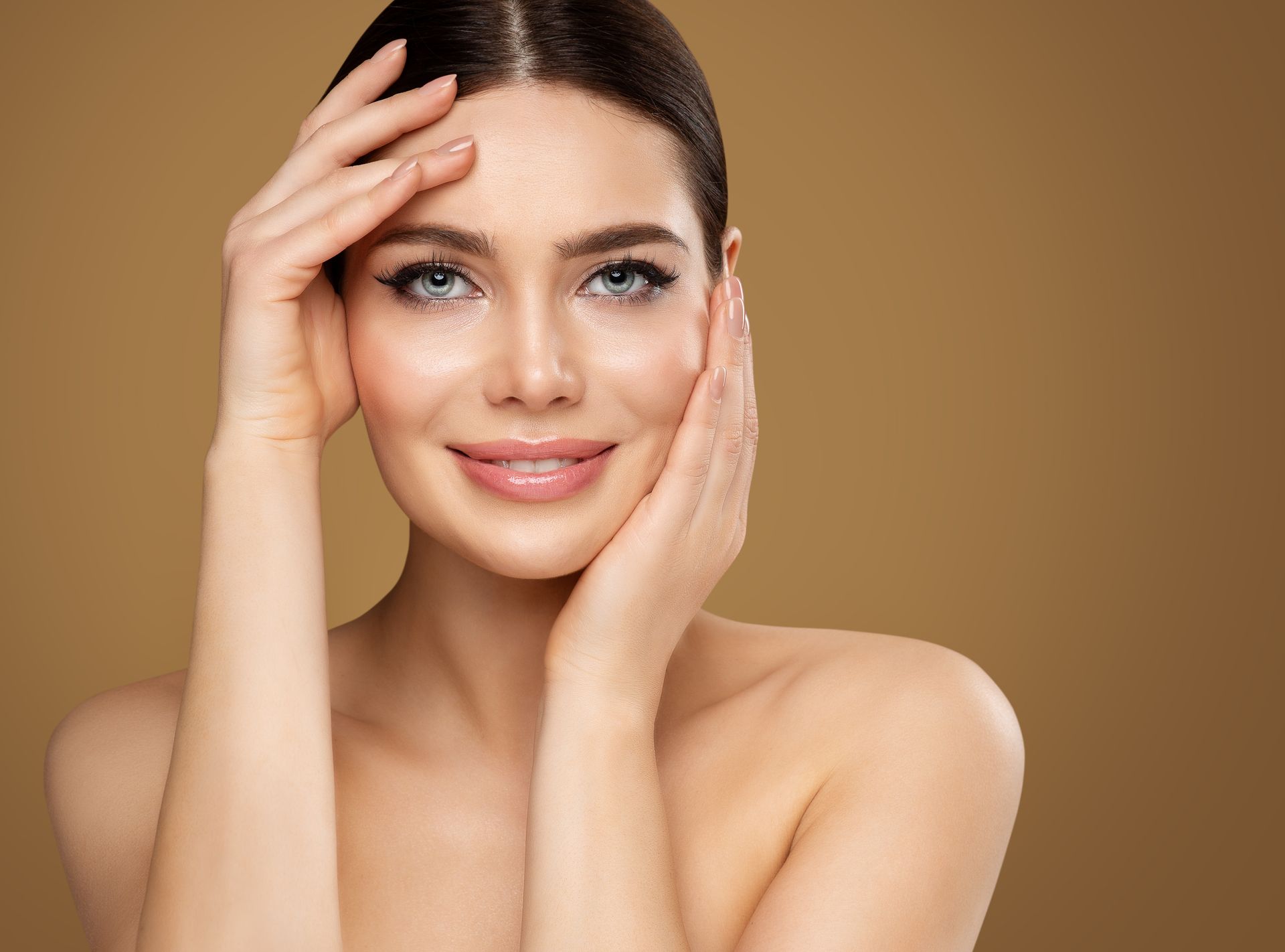 Woman posing against a plain background, touching her face with both hands.