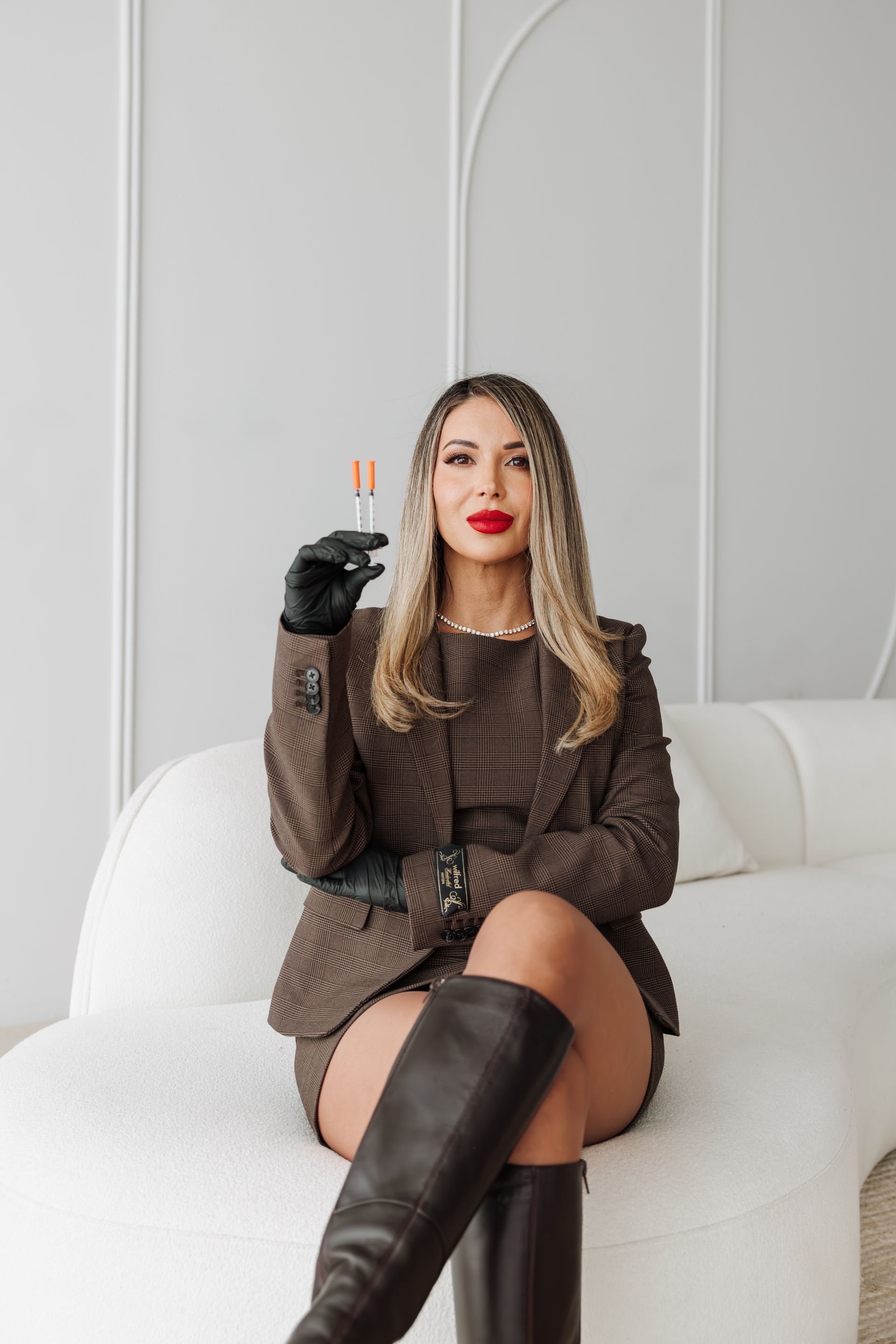 Woman in a brown outfit and black boots sitting on a white chair, holding a glass of champagne.