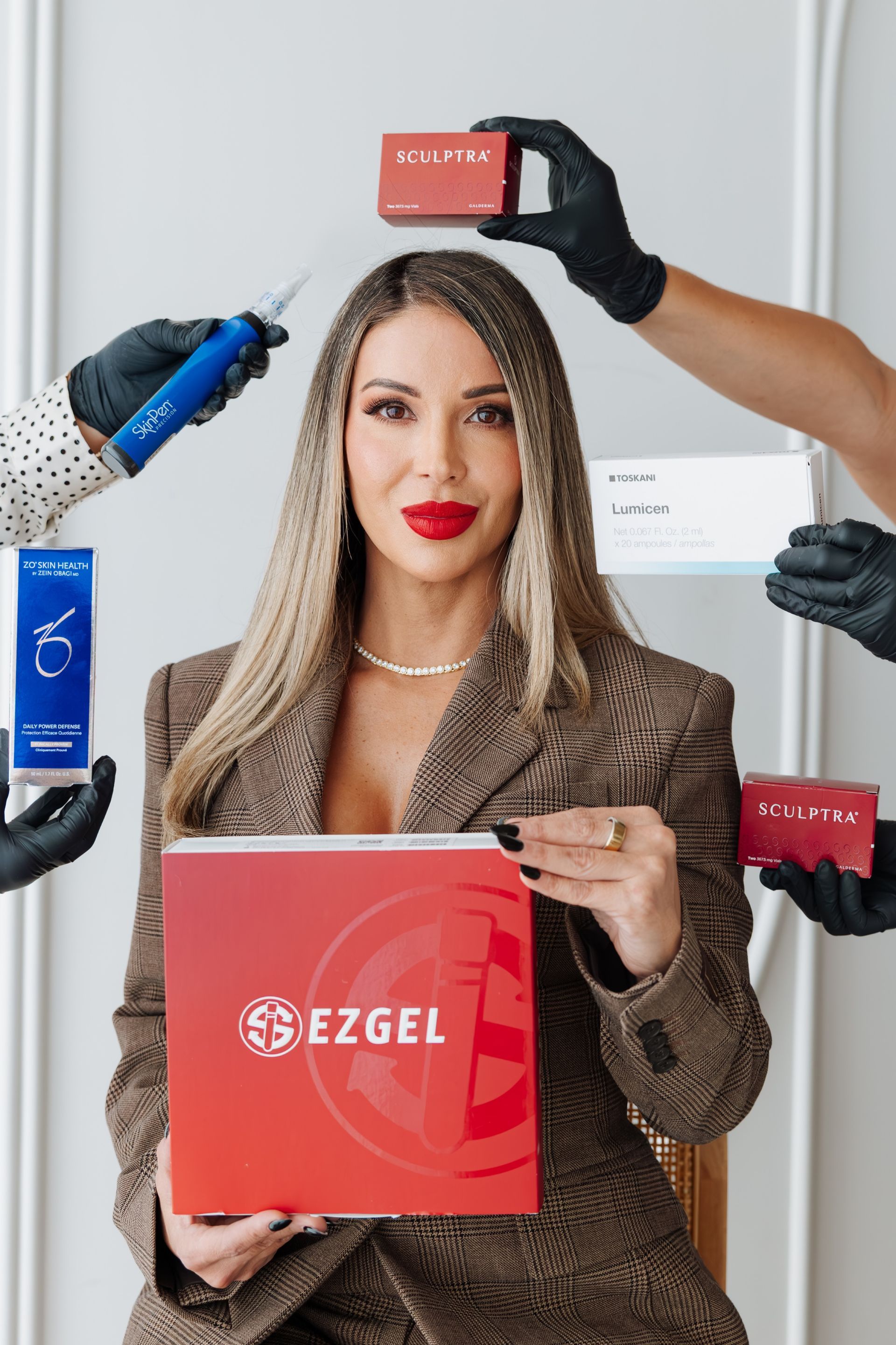 Woman posing with red EZGEL box while gloved hands hold beauty tools around her face in a studio setting