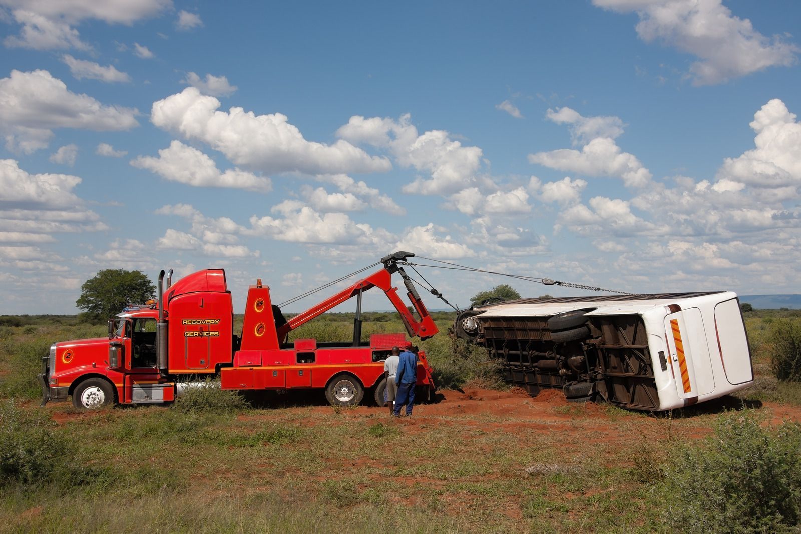 Red tow truck uprighting a white overturned bus on a grassy field under a cloudy blue sky.