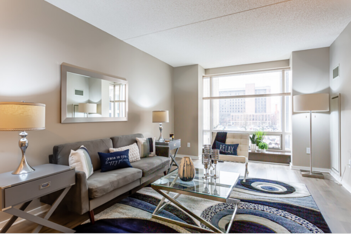 A modern living room with a gray couch, glass coffee table, and large window overlooking a city.