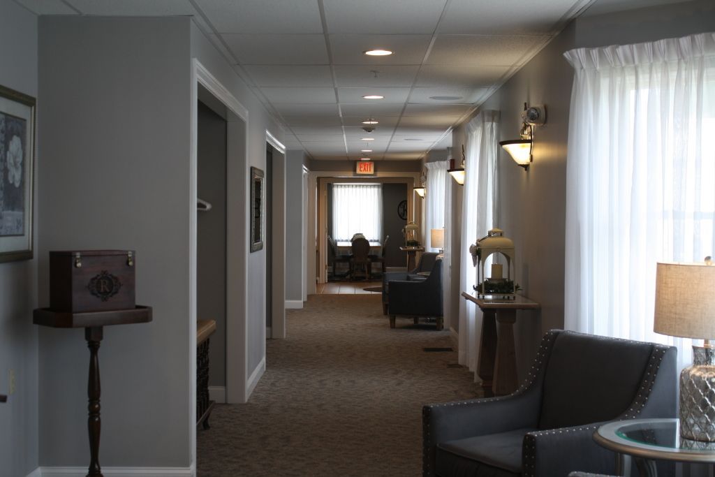 Carpeted hotel hallway with wall lights, chairs, and large windows at the end.