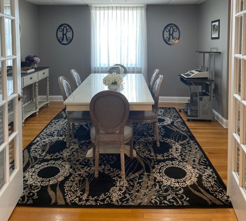 Formal dining room with white table, upholstered chairs, gray walls, and black patterned rug.