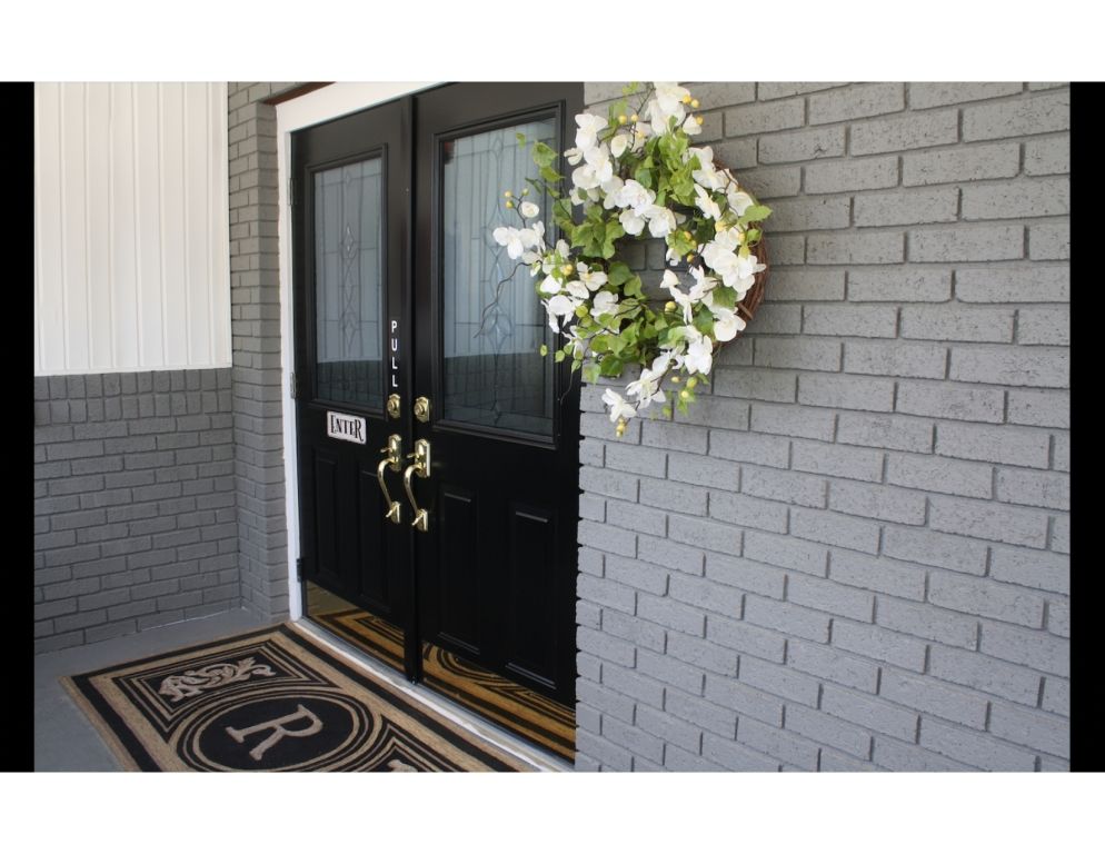 Black double front doors with a white floral wreath on a gray brick entryway, and a welcome mat.