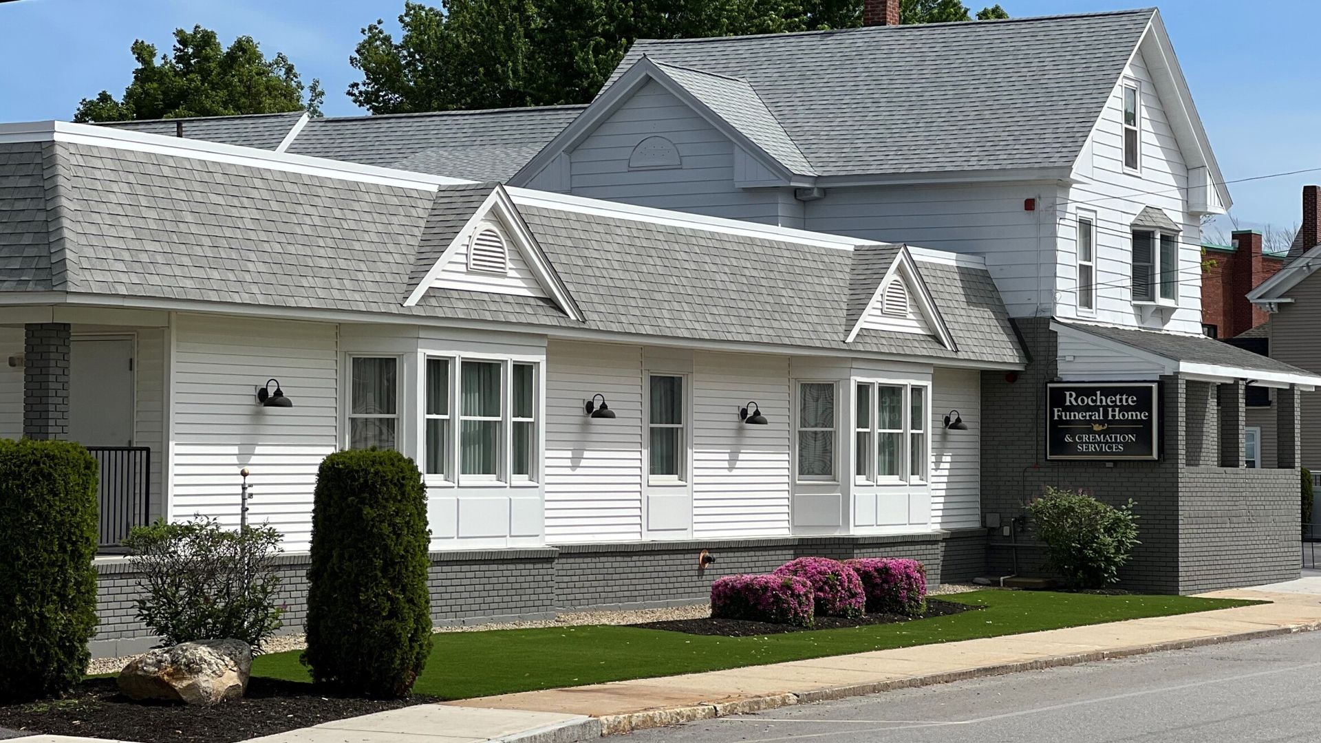 White roadside building with gray roof, small shrubs, and pink flowers in front