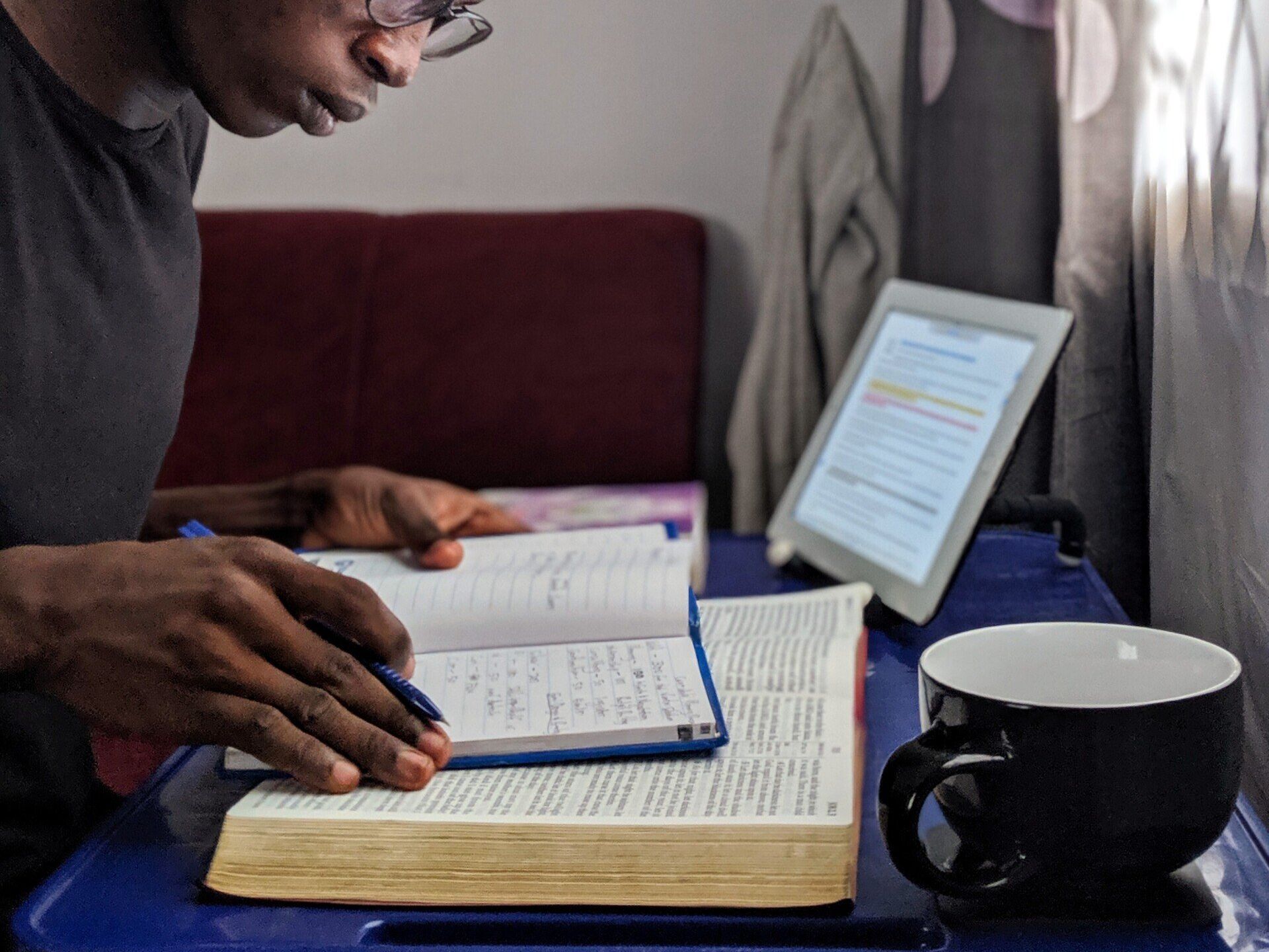 A man is sitting at a table reading a book and writing in a notebook.
