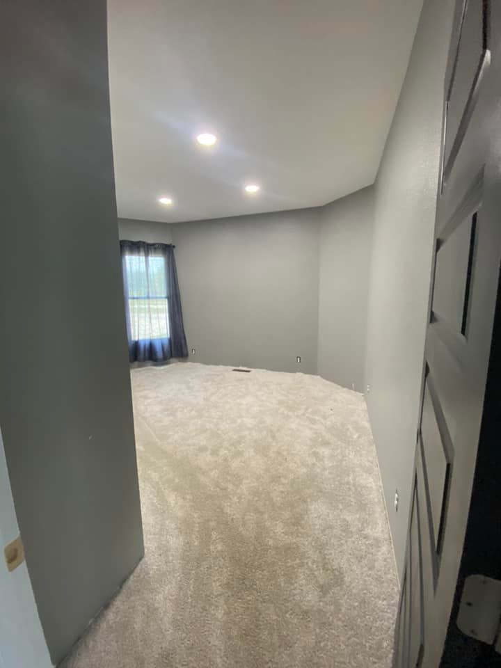 View from a doorway into an empty room with gray walls, beige carpet, and a window with dark curtains.