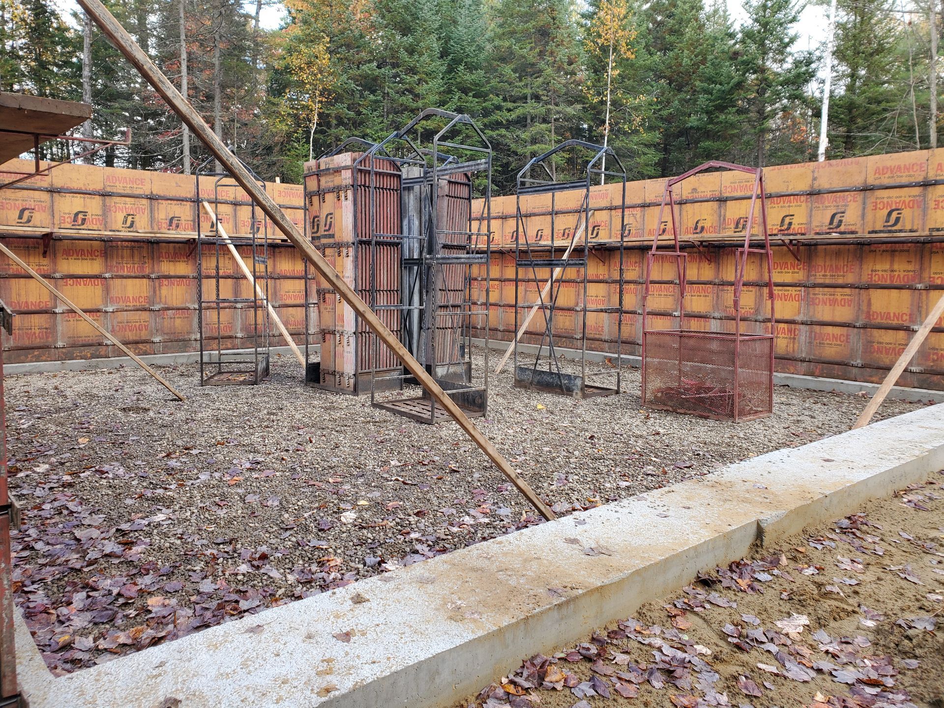 Vue extérieure d'une fondation en béton avec coffrages et armatures, posée sur du gravier et entourée d'arbres.