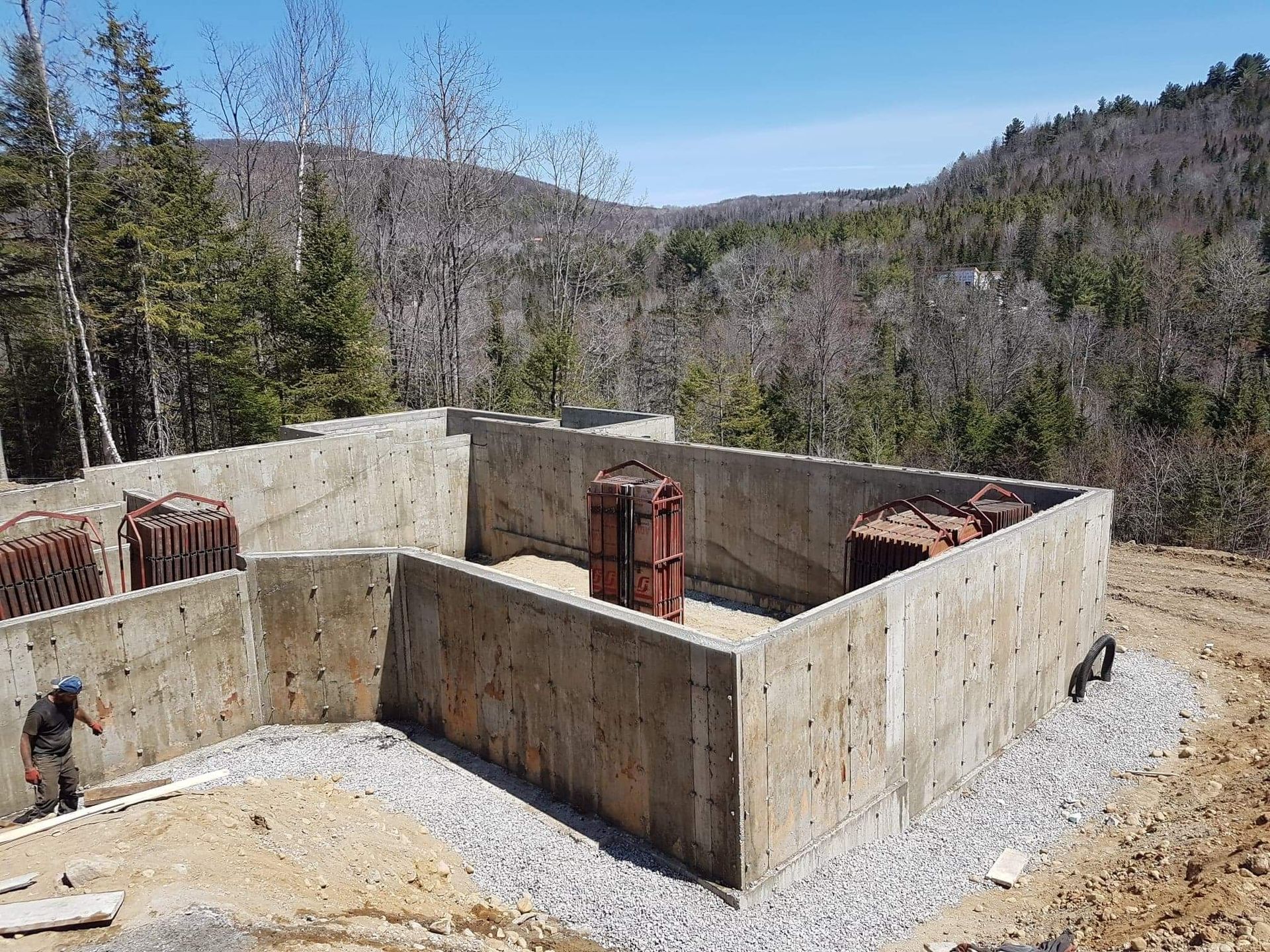 Fondations en béton avec structures métalliques ; une personne travaille à proximité ; arbres et collines en arrière-plan.