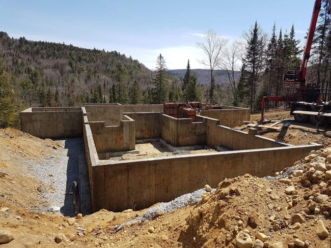 Fondations en béton en construction dans une zone boisée, sous un ciel dégagé, avec des engins de chantier.