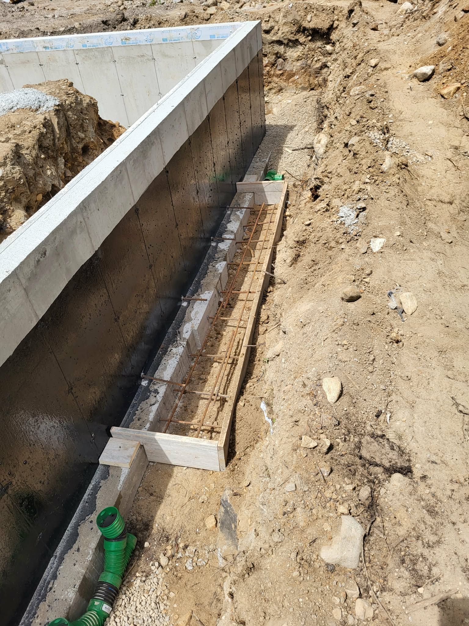 Chantier : mur de fondation en béton avec armatures apparentes et tuyaux de plomberie verts.