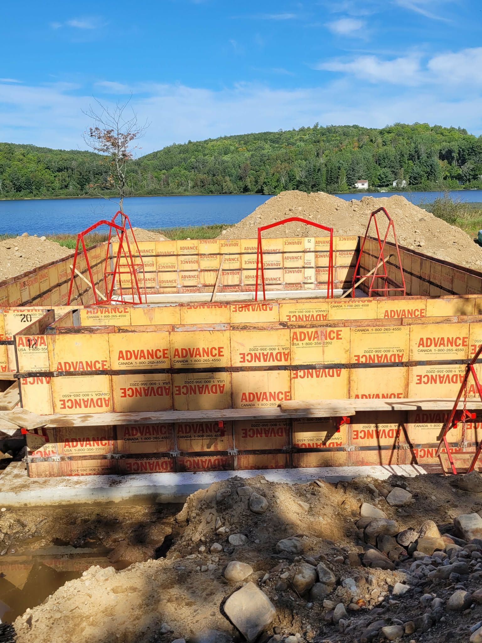 Chantier de construction avec coffrages en bois pour des fondations en béton, surplombant un lac arboré. Garde-corps de sécurité rouges.