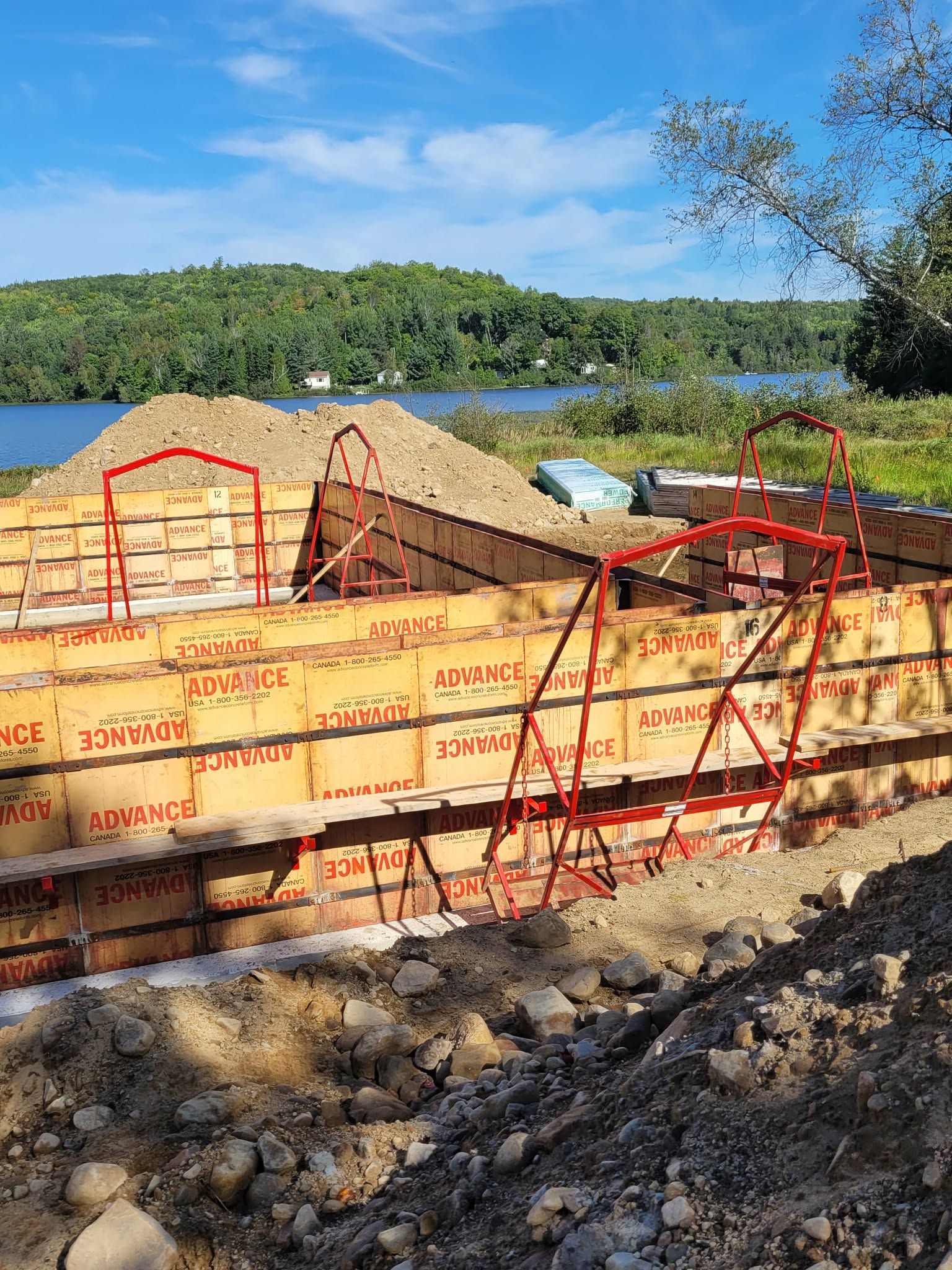Construction des fondations : coffrages bruns, poutres de soutien rouges, terre, lac et arbres en arrière-plan sous un ciel bleu.