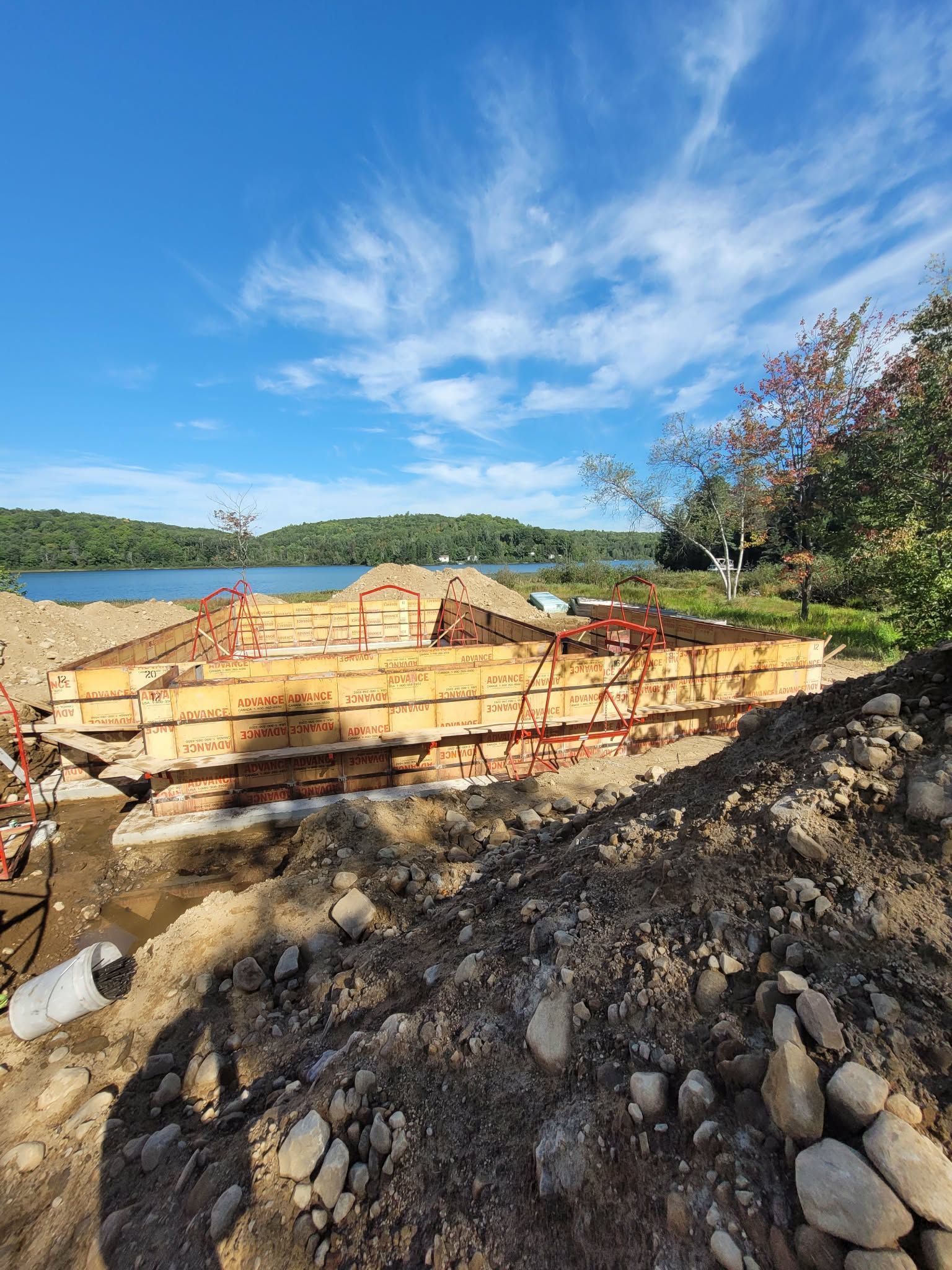 Chantier de construction avec coffrages en bois et terre, surplombant un lac et une rive boisée sous un ciel bleu.