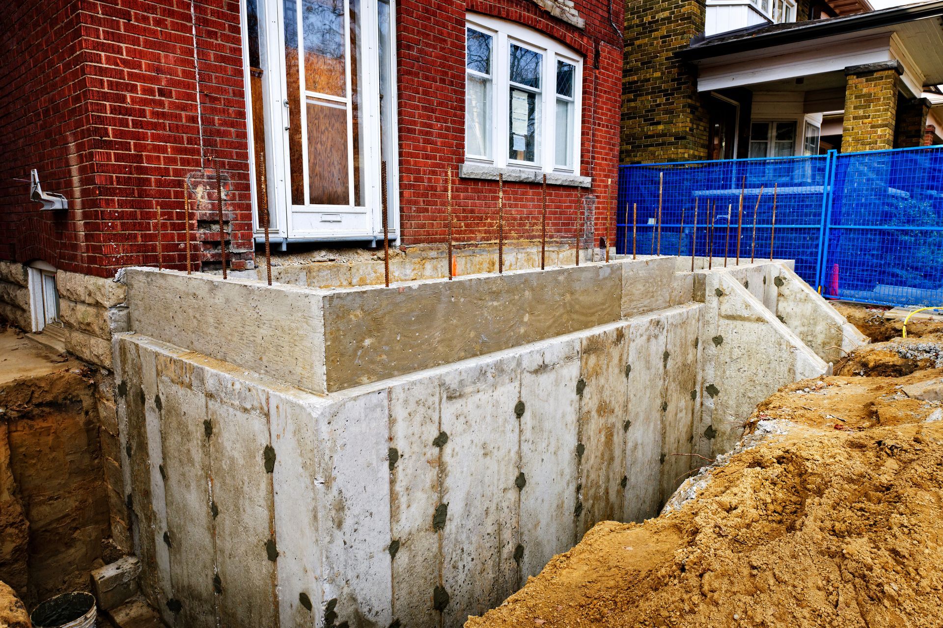 Fondations en béton nouvellement construites à côté d'une maison en briques rouges ; tas de terre et clôture bleue visibles.