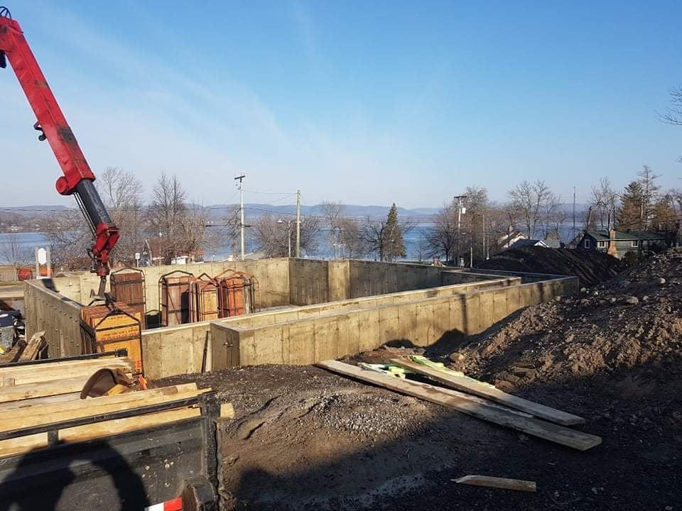 Chantier de construction avec fondations en béton et grue ; un lac et des arbres sont visibles en arrière-plan par une journée ensoleillée.