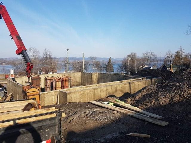 Chantier de construction avec fondations en béton et grue ; un lac et des arbres sont visibles en arrière-plan par une journée ensoleillée.