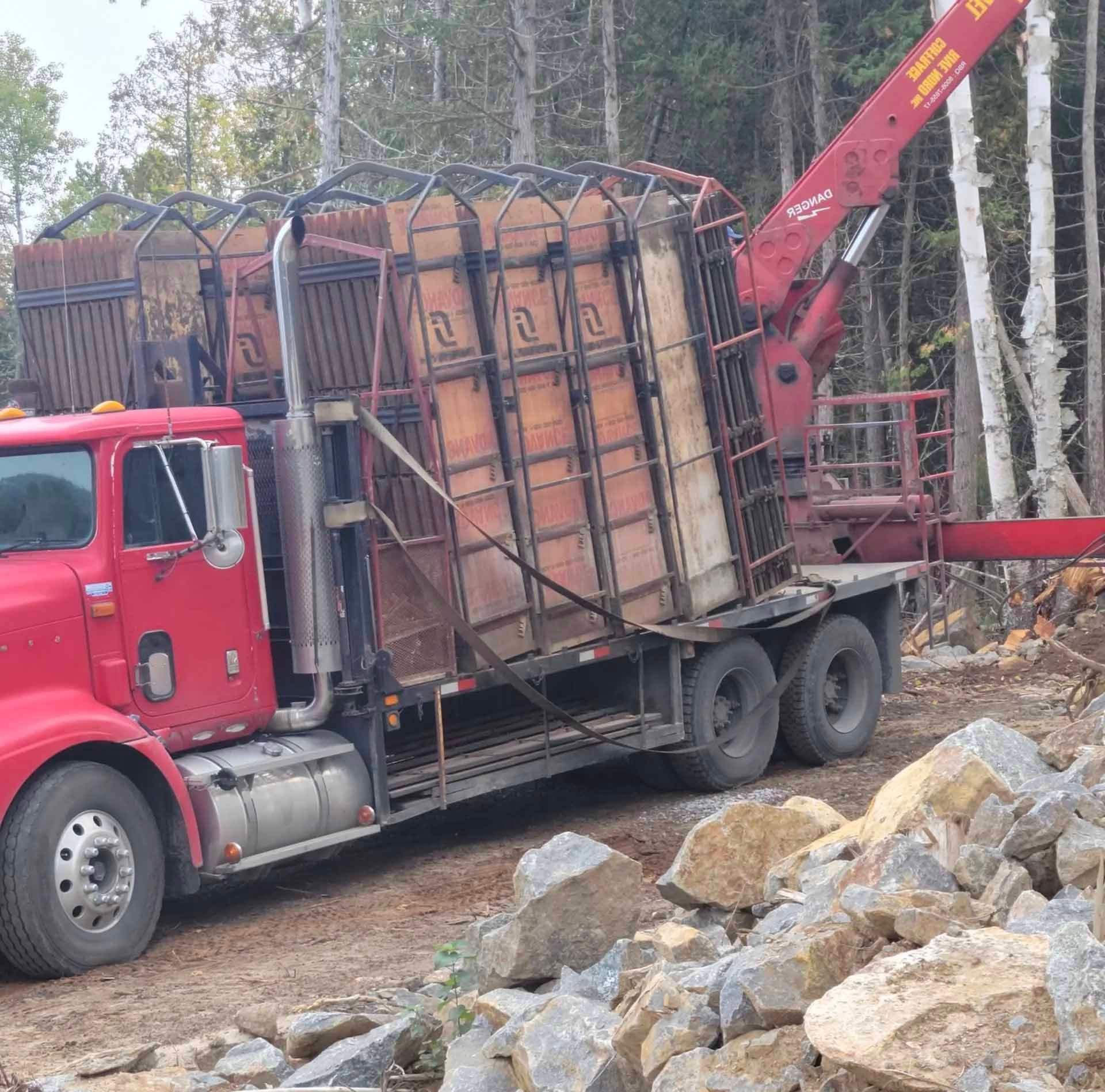 Un semi-remorque rouge équipé d'une grue transporte de grands conteneurs en bois sur un chemin de terre près de rochers et d'arbres.