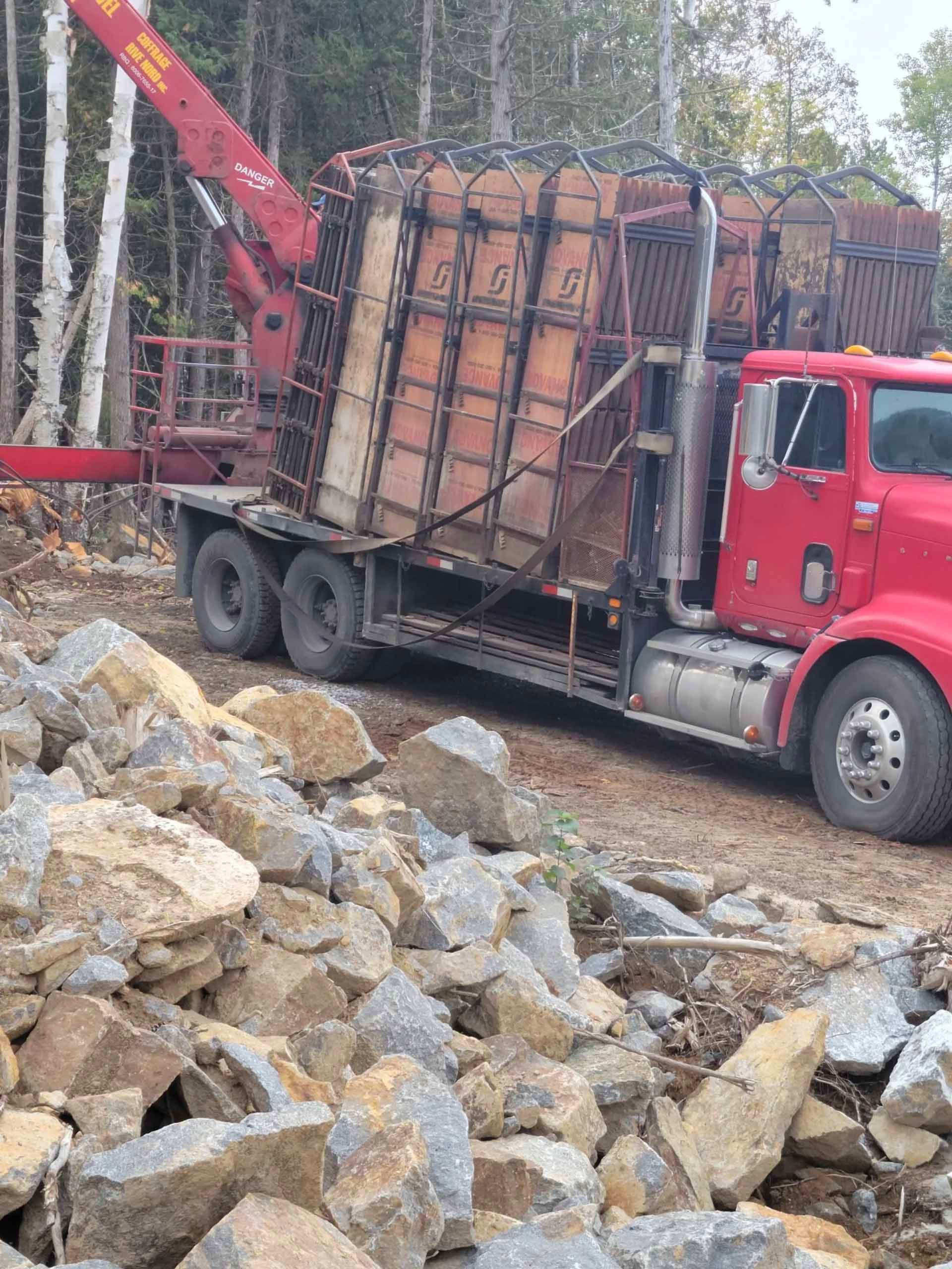 Camion rouge équipé d'une grue à flèche chargeant de grands conteneurs métalliques bruns remplis de pierres. Décor forestier.