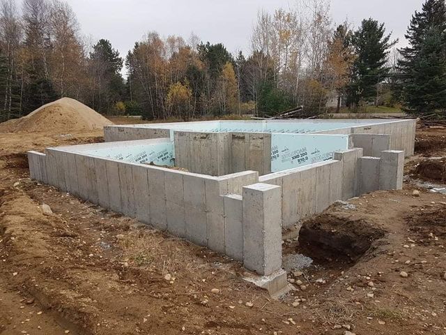 Fondations en béton d'un bâtiment en construction, entourées de terre et d'un tas de sable, avec des arbres en arrière-plan.