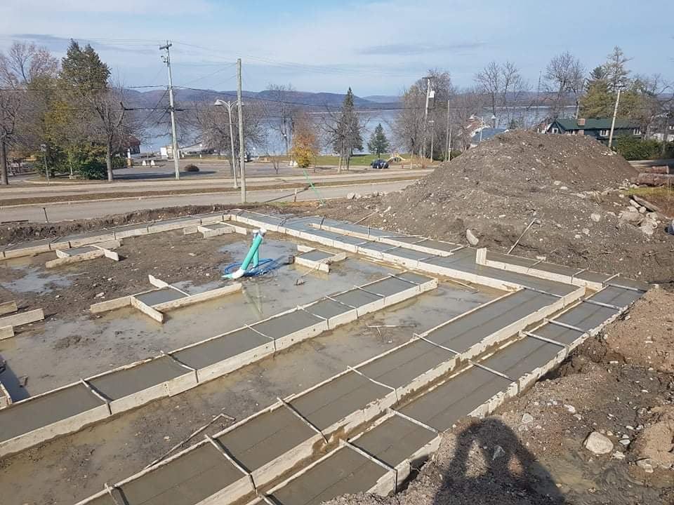 Chantier de construction avec murs de fondation en béton, tas de terre et vue sur un lac.