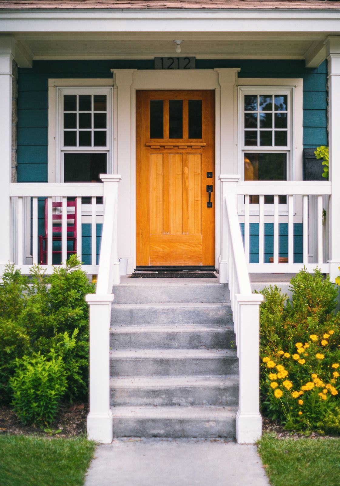 Front porch with orange door, white railings, gray steps, and green siding surrounded by shrubs and flowers