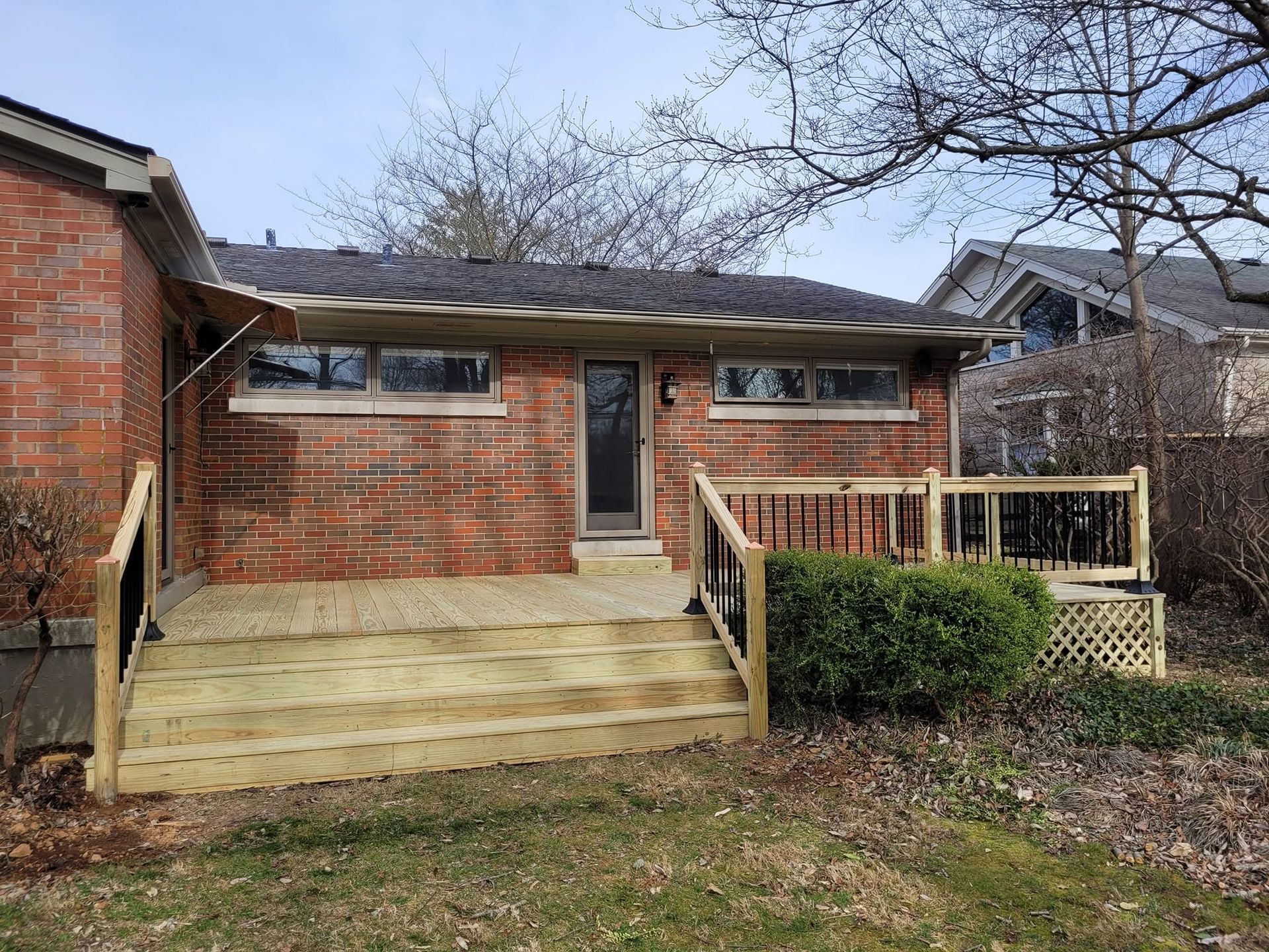 Brick house with a wooden front deck and steps, small shrubs, and leafless trees in the yard