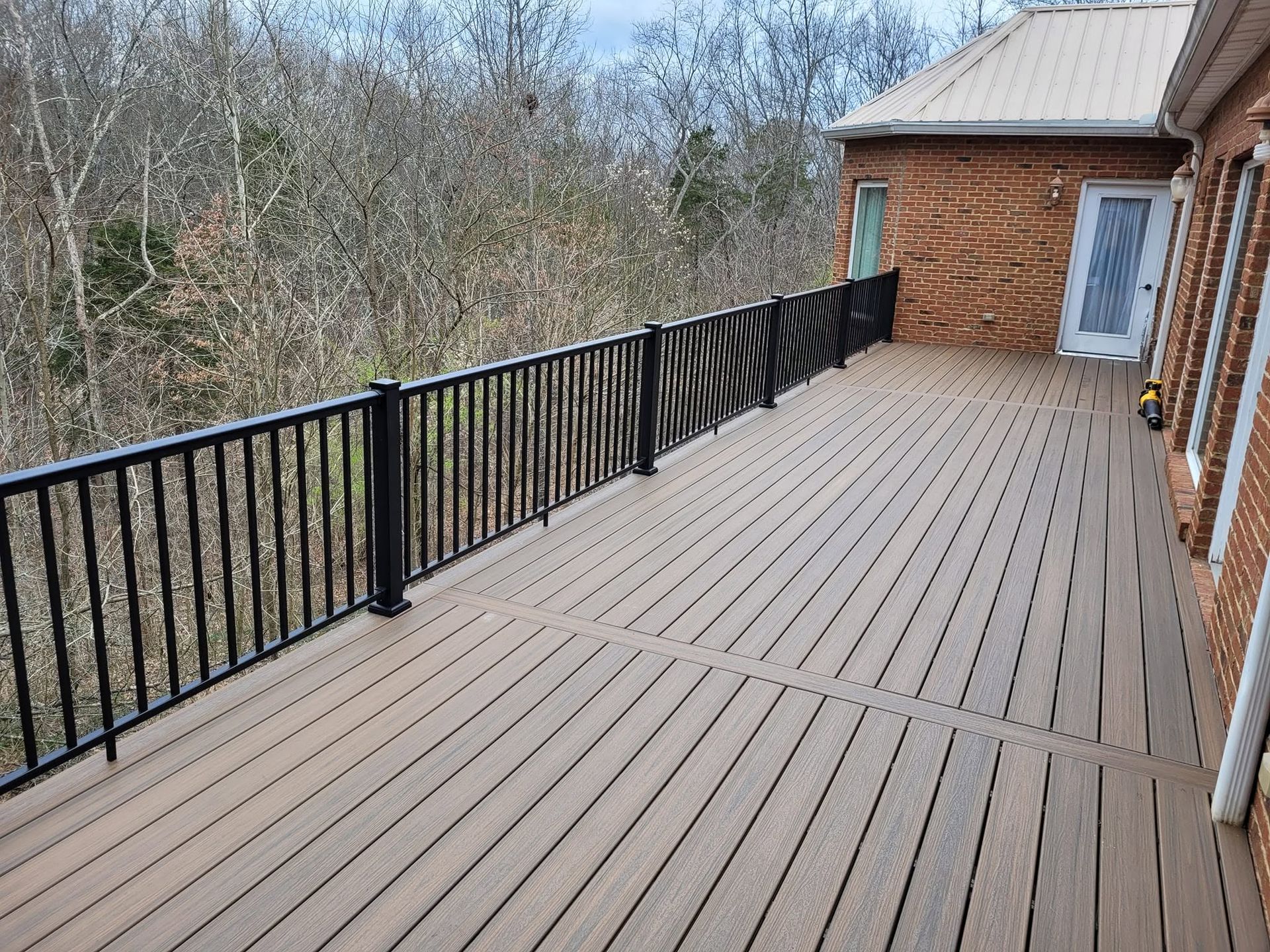 Long wooden deck with black railing beside a brick house, overlooking trees in a wooded area