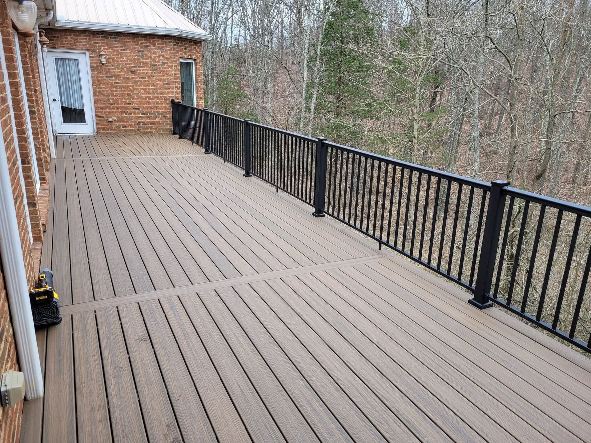 Empty wooden deck beside a brick house, with black railing and trees beyond