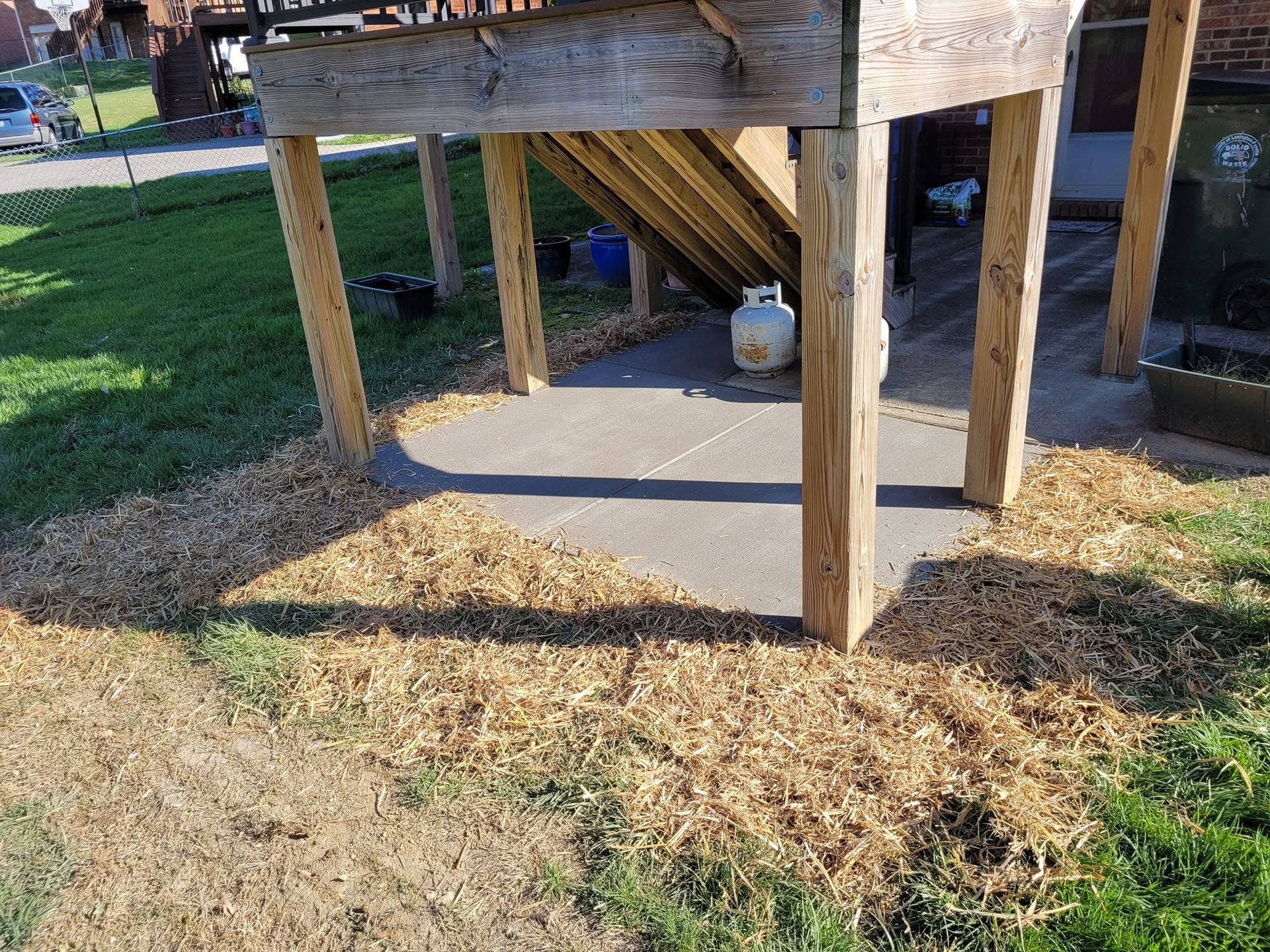 Wooden platform on legs over concrete, with a small white container underneath on a grassy outdoor area.
