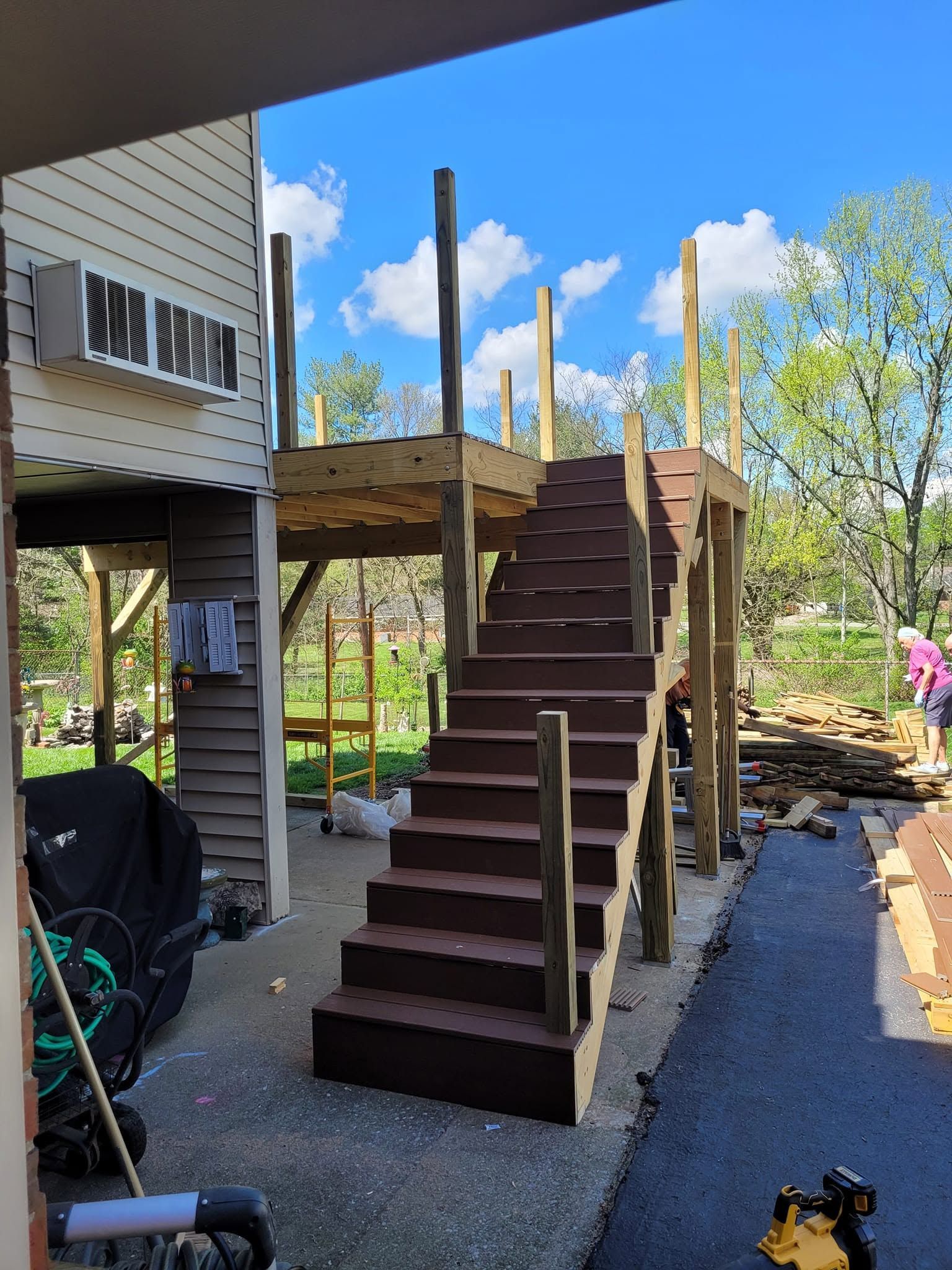Wooden stairs under construction beside a house, with railing posts and tools on a gravel yard.