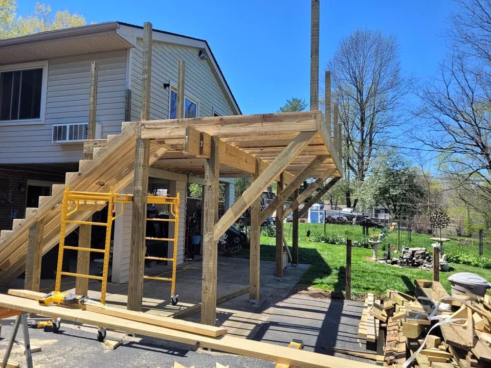 Wooden deck frame under construction beside a house, with ladders and stacked lumber in a yard.