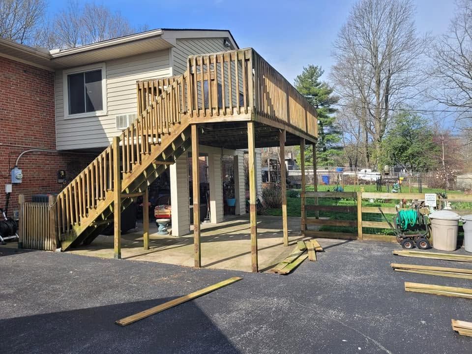 Wooden deck and staircase under construction beside a house, with lumber scattered on the driveway.